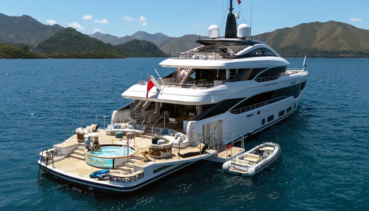 A luxury motor yacht with expansive deck space and a Jacuzzi at anchor in calm blue waters, with mountainous coastline in the background.